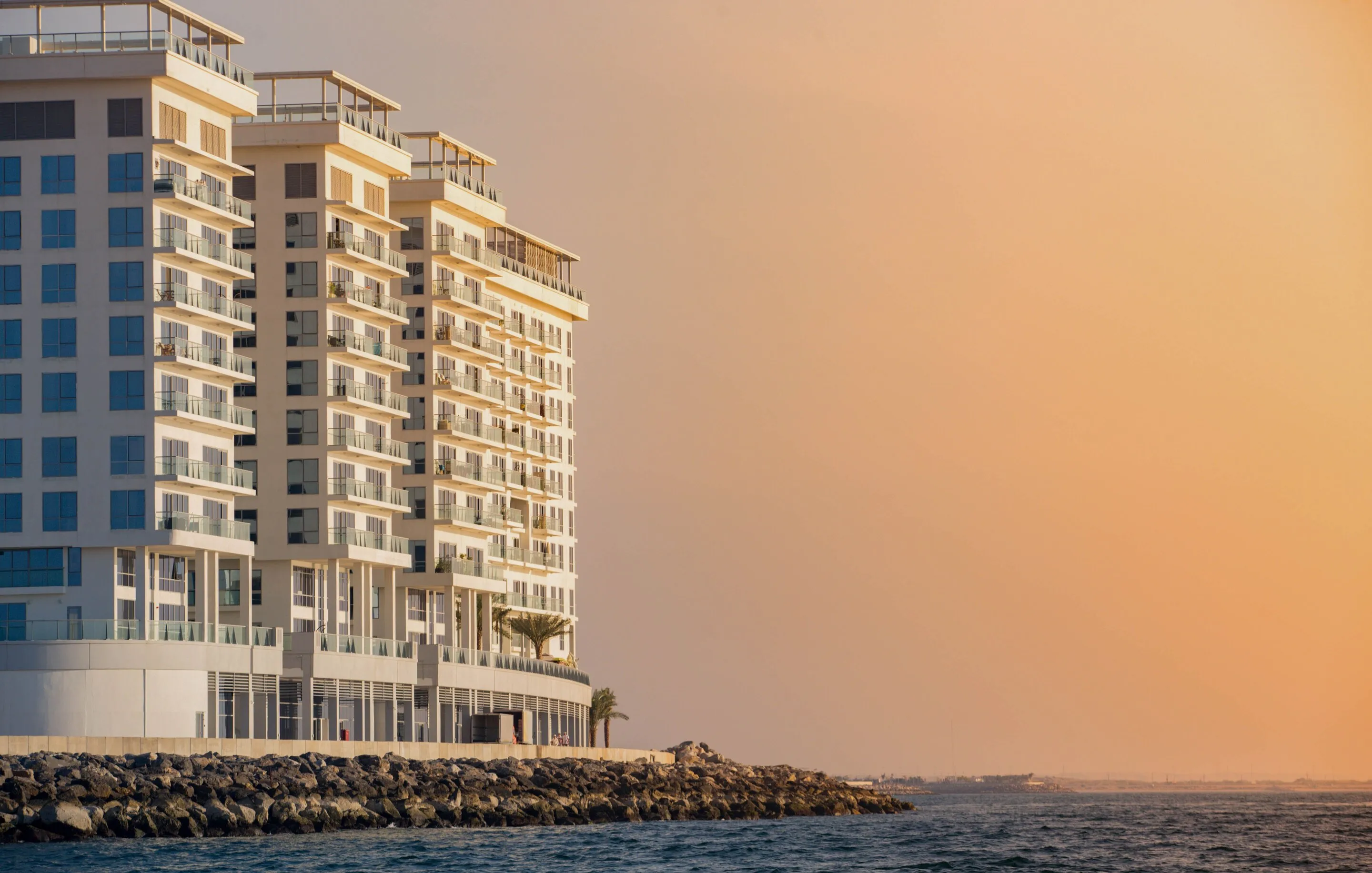 Modern white waterfront residential tower on a rocky sea point, glowing warm at golden-hour dusk over calm water
